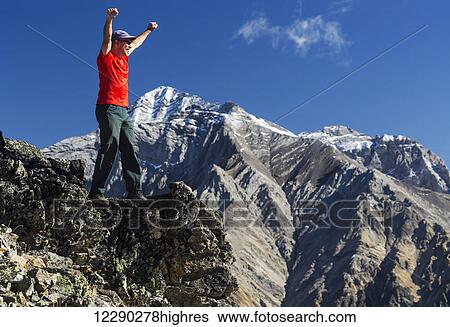 Stock Photograph - Male hiker standing with arms raised in the air on top of mountain ridge overlooking snow peaked mountains with blue sky and clouds, Kananaskis Provincial Park; Alberta, Canada. Fotosearch