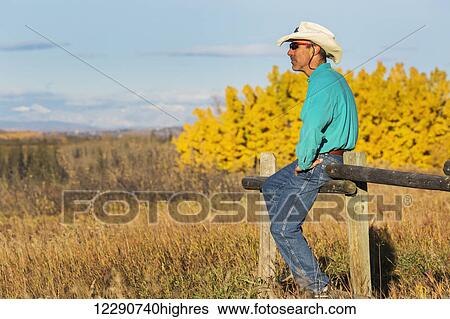 Stock Photograph - Male with straw cowboy hat and sunglasses sitting on top of a wooden fence looking into the distance with colourful trees in autumn in the background and blue sky; Calgary, Alberta, Canada. Fotosearch