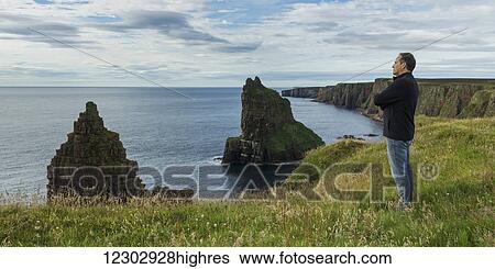 Stock Photograph - Man standing and looking out over the North Sea with rugged cliffs and sea stacks along the coastline; Scotland. Fotosearch
