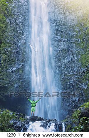 Stock Photograph - Man standing at the base of large waterfall in Hawaii. Fotosearch