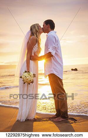 Stock Photograph - Married couple, bride and groom, kissing at sunset on beautiful tropical beach in Hawaii. Fotosearch
