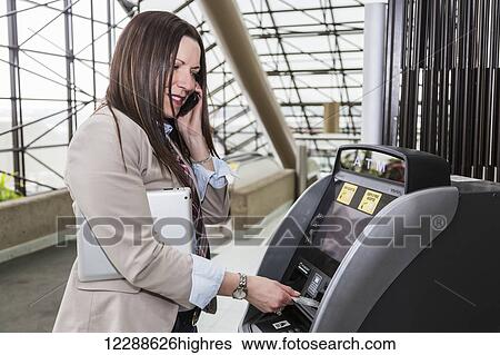 Stock Photograph - Mature business woman making a cash withdrawal at an ATM in the atrium of an office building; Edmonton, Alberta, Canada. Fotosearch