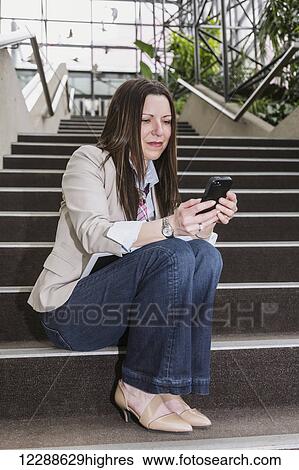 Stock Photograph - Mature business woman sitting on stairway in the atrium of an office building and texting on her smart phone; Edmonton, Alberta, Canada. Fotosearch