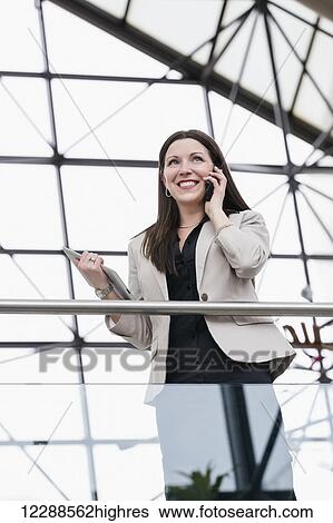 Stock Photograph - Mature business woman talking on her smart phone and holding a tablet in the atrium of an office building; Edmonton, Alberta, Canada. Fotosearch
