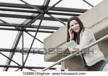 Stock Photograph - Mature business woman talking on her smart phone and holding a tablet in the atrium of an office building; Edmonton, Alberta, Canada. Fotosearch