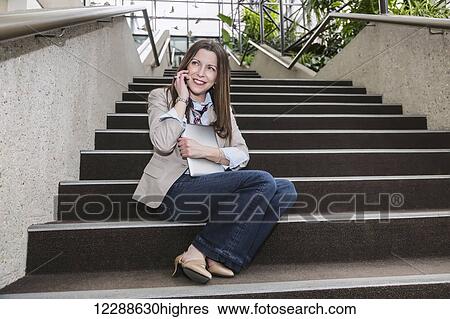 Stock Photograph - Mature business woman talking on her cell phone and holding a tablet while sitting on stairway in the atrium of an office building; Edmonton, Alberta, Canada. Fotosearch