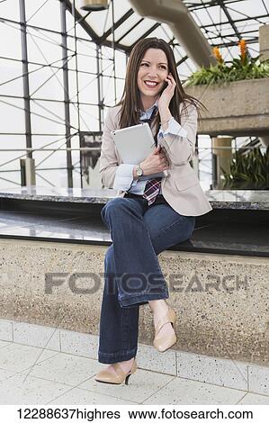 Stock Photograph - Mature business woman talking on her cell phone and holding a tablet in the atrium of an office building; Edmonton, Alberta, Canada. Fotosearch