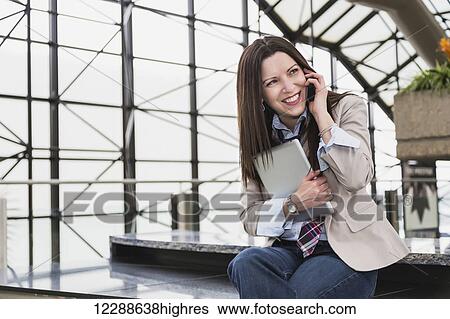 Stock Photograph - Mature business woman talking on her cell phone and holding a tablet in the atrium of an office building; Edmonton, Alberta, Canada. Fotosearch