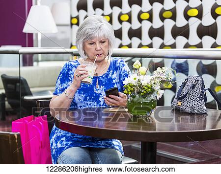 Stock Photograph - Mature woman checking her cell phone for messages while drinking an iced coffee at a cafe; St. Albert, Alberta, Canada. Fotosearch