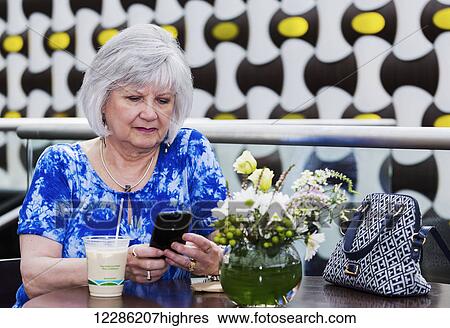 Stock Photograph - Mature woman checking her cell phone for messages while drinking an iced coffee at a cafe; St. Albert, Alberta, Canada. Fotosearch