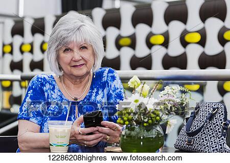 Stock Photograph - Mature woman checking her cell phone for messages while drinking an iced coffee at a cafe; St. Albert, Alberta, Canada. Fotosearch