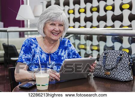 Stock Photograph - Mature woman using her tablet while taking a coffee break at a cafeteria; St. Albert, Alberta, Canada. Fotosearch