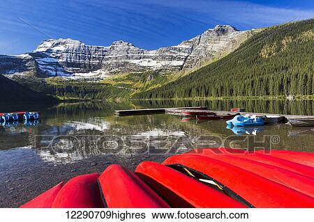 Stock Photograph - Mountain lake with dock reflecting snow peaks and blue sky with upside down red canoes in the foreground; Waterton, Alberta, Canada. Fotosearch