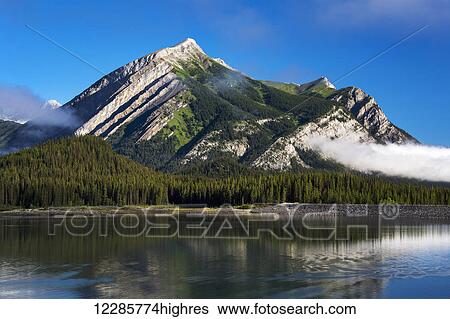 Mountain reflecting in lake with fog patches and blue sky; Kananaskis Country, Alberta, Canada View Large Photo Image Stock Photograph - Mountain reflecting in lake with fog patches and blue sky; Kananaskis Country, Alberta, Canada. Fotosearch