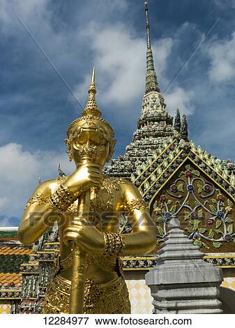 Stock Photo - Ornate building with spire and a gold statue in the foreground, Temple of the Emerald Buddha (Wat Phra Kaew); Bangkok, Thailand. Fotosearch