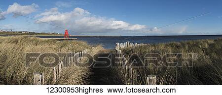 Stock Photograph - Path lined with wooden posts and tall grass at the coast, Herd Groyne Lighthouse in the distance; South Shields, Tyne and Wear, England. Fotosearch