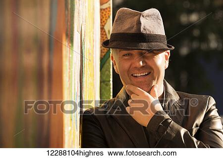 Stock Photograph - Portrait of a smiling man in a leather jacket and grey hat with his hand on his chin, standing against a sunlit wall with a colourful mural on it; London, England. Fotosearch