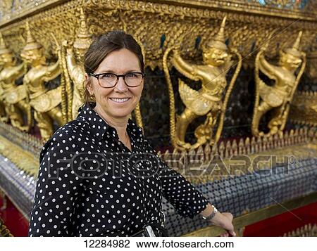 Stock Image - Portrait of a woman standing beside a wall with gold statues, Temple of the Emerald Buddha (Wat Phra Kaew); Bangkok, Thailand. Fotosearch