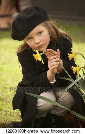 Portrait of a young girl dressed in a brown overcoat, trousers and beret holding a yellow daffodil with one hand and stroking it with the other, crouching on grass and looking serious; London, England View Large Photo Image Stock Photograph - Portrait of a young girl dressed in a brown overcoat, trousers and beret holding a yellow daffodil with one hand and stroking it with the other, crouching on grass and looking serious; London, England. Fotosearch