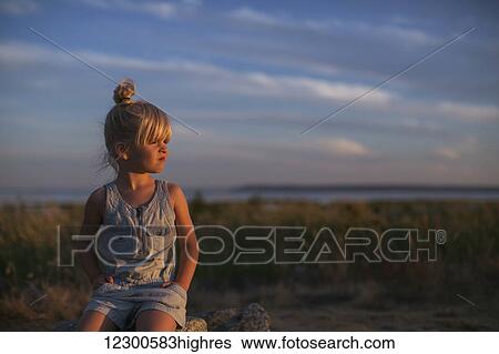 Stock Photograph - Portrait of a young girl sitting on a rock on a beach at sunset; Surrey, British Columbia, Canada. Fotosearch