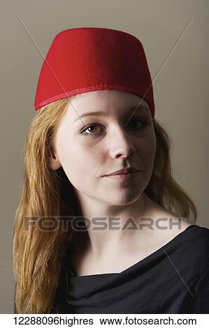Stock Photograph - Portrait of a young woman with long red hair wearing a red felt Moroccan fez; Caldecott, England. Fotosearch