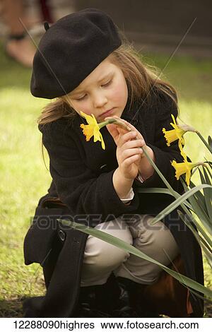 Portrait of young girl dressed in a brown overcoat, trousers and beret is holding a yellow daffodil with one hand and stroking it with the other, crouching on the grass, staring at the daffodil and looking sad; London, England View Large Photo Image Stock Photograph - Portrait of young girl dressed in a brown overcoat, trousers and beret is holding a yellow daffodil with one hand and stroking it with the other, crouching on the grass, staring at the daffodil and looking sad; London, England. Fotosearch