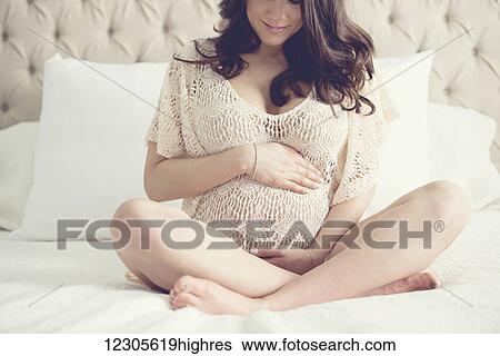 Stock Photograph - Pregnant woman sitting cross-legged on bed with hands on top of and underneath stomach; Toronto, Ontario, Canada. Fotosearch
