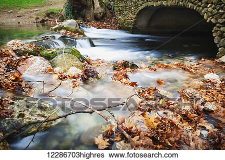 Stock Photograph - River flowing under stone bridge with floating autumn coloured leaves; Naoussa, Greece. Fotosearch