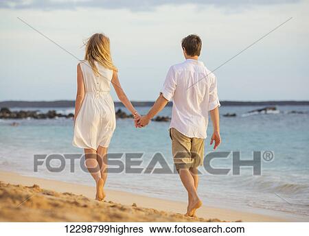 Stock Photograph - Romantic happy couple walking on beach at sunset. Smiling holding hands. Man and woman in love. Fotosearch