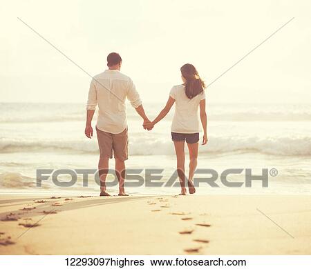 Stock Photograph - Romantic Young Lovers Walking Down the Beach at Sunset on Vacation. Fotosearch