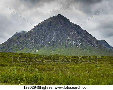 Stock Photograph - Rugged peaked mountain under a cloudy sky with lush grass in a field; Scotland. Fotosearch