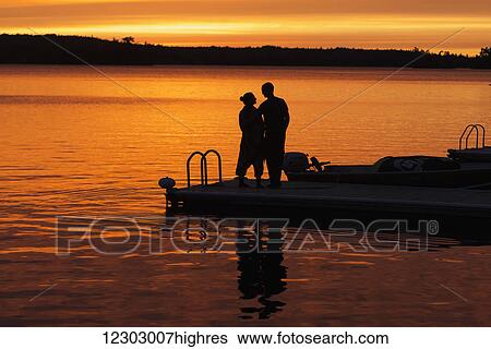 Silhouette of a couple standing at the end of a dock at sunset with an orange glow in the sky and reflected in the tranquil lake water; Ontario, Canada View Large Photo Image Stock Photograph - Silhouette of a couple standing at the end of a dock at sunset with an orange glow in the sky and reflected in the tranquil lake water; Ontario, Canada. Fotosearch