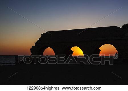 Stock Photograph - Silhouette of a Roman aqueduct with arches at sunset in Caesarea Maritima National Park; Israel. Fotosearch