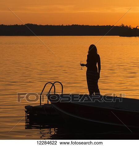 Stock Image - Silhouette of a woman holding a glass of wine while standing on a dock on a golden lake at sunset; Lake of the Woods, Ontario, Canada. Fotosearch