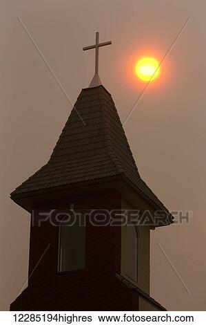 Stock Photograph - Silhouette of church steeple with orange sun ball in a hazy sky; Calgary, Alberta, Canada. Fotosearch
