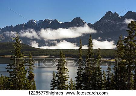 Stock Photograph - Silhouette of mountain range with low cloud in valley along mountain lake with blue sky; Kananaskis Country, Alberta, Canada. Fotosearch
