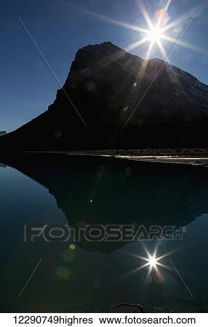 Stock Photograph - Silhouette of mountain reflecting in lake with sun burst and blue sky, Banff National Park; Alberta, Canada. Fotosearch