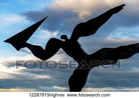Stock Photograph - Silhouetted sculpture of a person about to throw a paper aeroplane, called Utkast, by Kare Groven, at Gardermoen Airport against a mixture of grey clouds and blue sky just after sunset; Akershus, Norway. Fotosearch