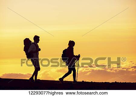 Stock Photograph - Silhouettes of two hikers with backpacks walking at sunset. Trekking and enjoying the sunset view from mountain top above the clouds.. Fotosearch