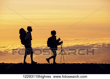 Stock Photograph - Silhouettes of two hikers with backpacks walking at sunset. Trekking and enjoying the sunset view from mountain top above the clouds.. Fotosearch