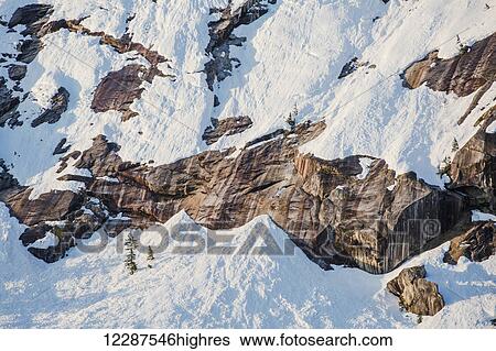 Stock Photograph - Snow covered cliffs and evergreen trees bathed in sunset light on the shore of Kings Bay, Prince William Sound; Alaska, United States of America. Fotosearch