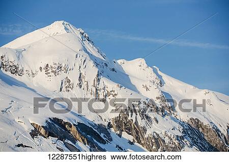 Stock Photograph - Snow covered cliffs bathed in sunset light on the shore of Kings Bay, Prince William Sound; Whittier, Alaska, United States of America. Fotosearch