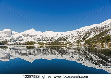 Stock Photograph - Snow covered mountain peaks reflect off calm waters on a clear day in Kings Bay, Prince William Sound; Whittier, Alaska, United States of America. Fotosearch