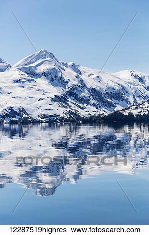 Stock Photograph - Snow covered mountains reflect in the calm waters of Prince William Sound in winter, Kings Bay; Alaska, United States of America. Fotosearch