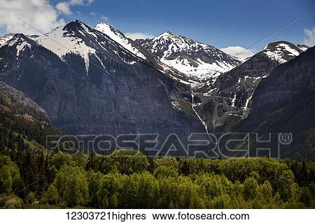 Stock Photograph - Snowcapped Telluride, Colorado mountains with a waterfall and stream tumbling down a steep mountainside and sunlit trees in the foreground; Colorado, United States of America. Fotosearch