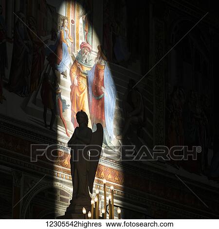 Stock Photograph - Statue of an angel in front of partly illuminated artwork on a wall; Florence, Italy. Fotosearch