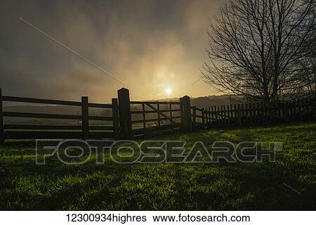 Stock Photograph - Sunlight shining through the clouds and a shadow of a fence cast on the grass; Beamish, England. Fotosearch