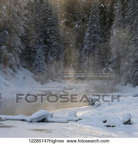Stock Photograph - Sunlight through the misty air over a snow covered landscape and bridge crossing over water; Emerald Lake, British Columbia, Canada. Fotosearch