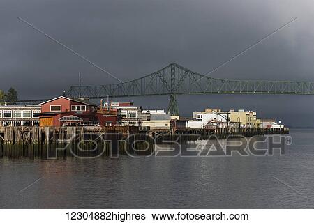 Stock Photograph - Sunlight warms the riverfront with storm clouds over the Astoria-Megler bridge; Astoria, Oregon, United States of America. Fotosearch
