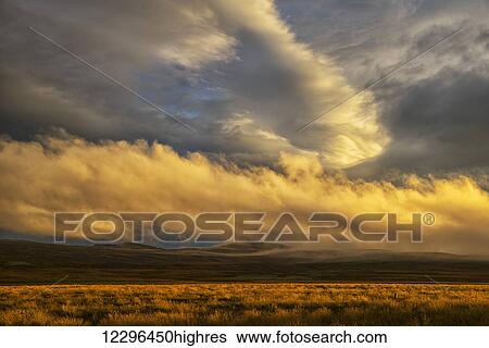 Stock Photograph - Sunset lights up the clouds while late day sun casts a warm glow on the grass; Iceland. Fotosearch